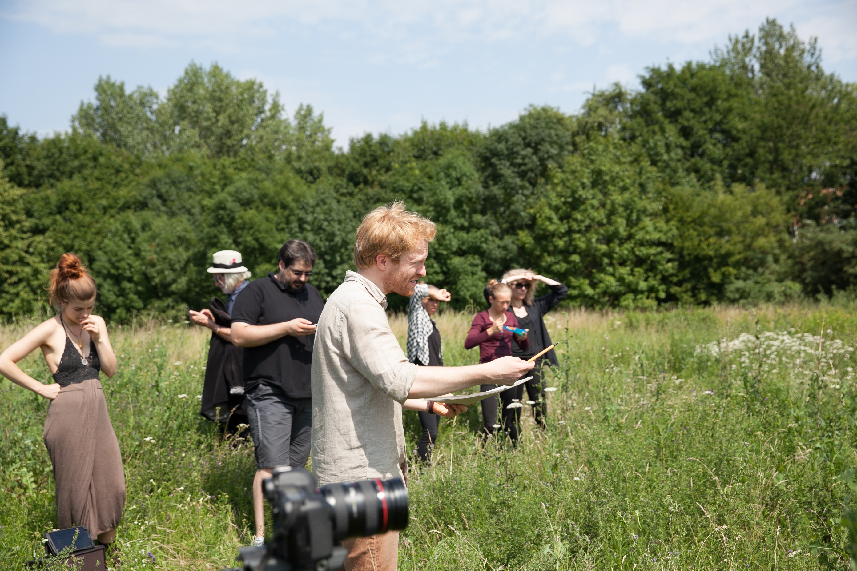 Outdoor film production: Oliver Hasse is seen in side profile, directing. Camera in the foreground, cast in the background. High grass everywhere, green trees and a blue sky.